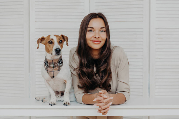 Photo of good looking woman with dark hair, spends leisure time with jack russel terrier dog, loves animals, have friendly relationship with pet, pose together at white table, play together. - Photo, image