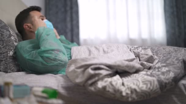 Kaszel mężczyzna w kombinezonie ochronnym wprowadzenie maski twarzy leżącej w łóżku obok szafki nocnej z wielu leków i pigułek - Materiał filmowy, wideo