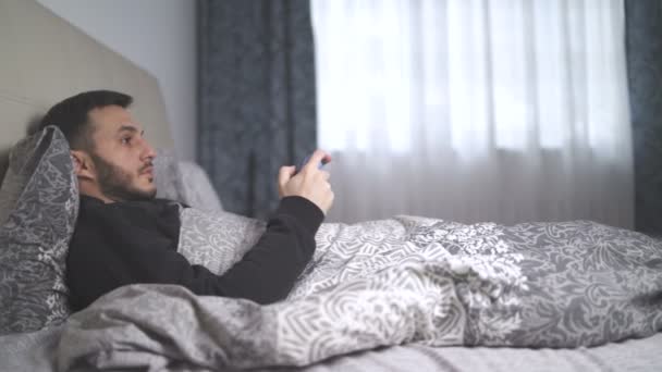 Chory młodzieniec grający w gry wideo z łóżka. Mężczyzna kaszle i leży w łóżku - Materiał filmowy, wideo