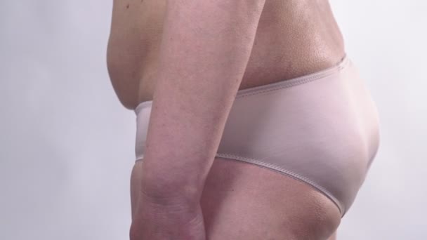 Een vrouw van middelbare leeftijd met een slappe huid draait naar de zijkanten en toont haar dikke buik, op een witte achtergrond. Zijplan. - Video