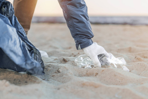 Close up of man's hand picking up trash and plastics cleaning the beach with a garbage bag - Фото, изображение