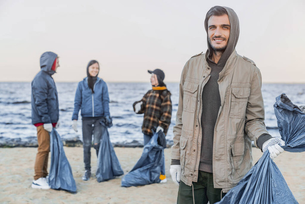 Porträt eines jungen männlichen Freiwilligen gemischter Rasse, der in die Kamera lächelt, nachdem er mit seinen Freunden Plastikmüll am Strand aufgesammelt hat - Foto, Bild