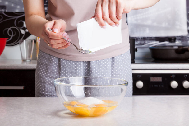 Τα γυναικεία χέρια κρατούν στα χέρια τους μια σακούλα βανίλιας και τη χύνουν σε ένα κουτάλι για να την προσθέσουν στη ζύμη ζύμωσης - χτυπημένα αυγά με αλεύρι και ζάχαρη στο φόντο των μαγειρικών σκευών. Μαγειρική πίτα στο σπίτι - Φωτογραφία, εικόνα