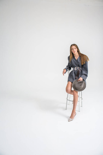 An attractive young girl, a model who looks to the side and straightens her hair in a gray jacket and hat, is sitting on a chair. Men's style clothing. isolated on a white background. Selective focus - Foto, Imagem