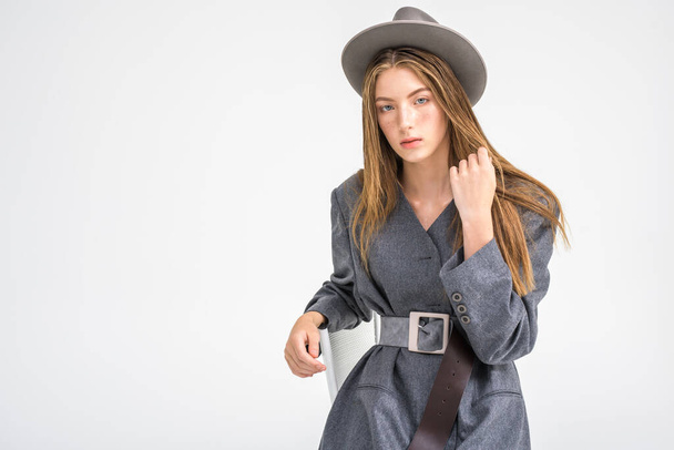 An attractive young girl, a model who looks to the side and straightens her hair in a gray jacket and hat, is sitting on a chair. Men's style clothing. isolated on a white background. Selective focus - Φωτογραφία, εικόνα