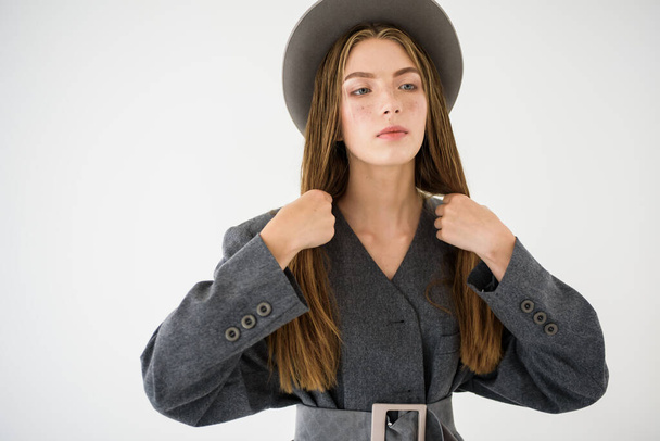 Vonzó fiatal lány, egy modell, aki oldalra néz, kiegyenesíti a haját szürke kabátban, és kalapot tart a kezében. Férfias ruha. Elszigetelve, fehér háttérrel. Szelektív fókusz - Fotó, kép