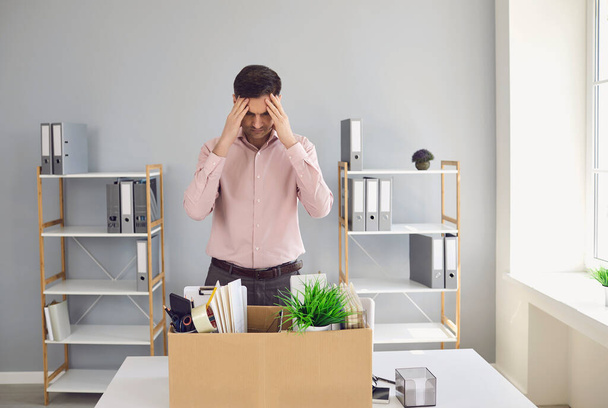 Unemployment. Dismissal. Dismissed businessman is upset with a cardboard box holding his head in his hands while sitting in a company office. - Fotografie, Obrázek