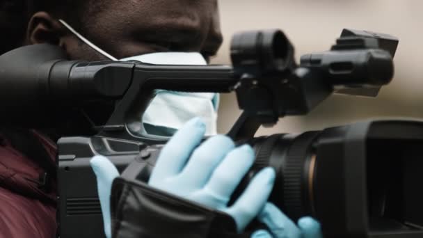 Afrikanischer Kameramann mit medizinischer Maske hält die Kamera mit den Latexhandschuhen. Berichterstattung über Coronavirus-Pandemie.  - Filmmaterial, Video