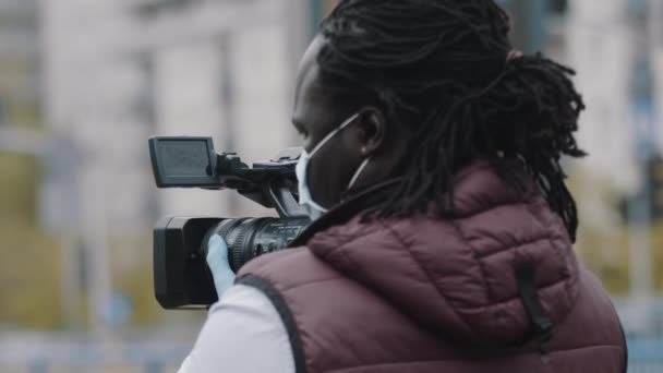 Tıbbi maskeli Afrikalı muhabir lateks eldivenli kamerayı tutuyor. Coronavirüs salgınını bildiriyorum. - Video, Çekim