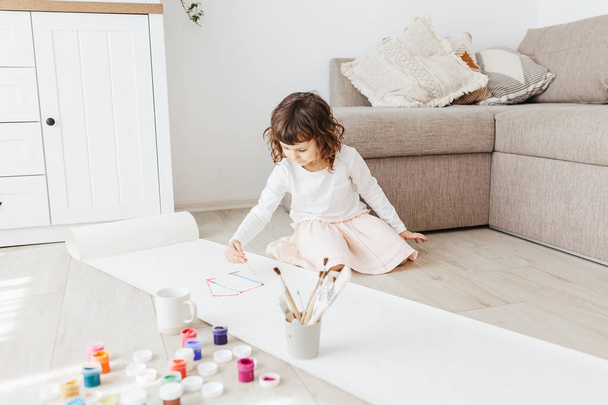Cute happy little girl, adorable preschooler, painting house with paints on paper roll sitting on floor in a sunny white living room at home, creative young artist at work - Photo, Image