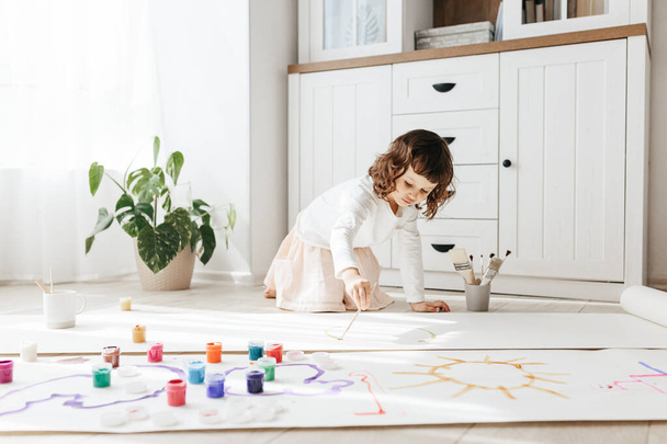 Cute happy little girl, adorable preschooler, painting with paints on paper roll sitting on floor in a sunny white living room at home, creative young artist at work - Photo, Image