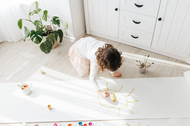 Roztomilá šťastná holčička, rozkošný předškolák, malování slunce s barvami na papíře role sedí na podlaze v slunném bílém obývacím pokoji doma, kreativní mladý umělec v práci - Fotografie, Obrázek