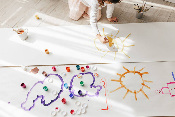 Cute happy little girl, adorable preschooler, painting sun with paints on paper roll sitting on floor in a sunny white living room at home, creative young artist at work - Photo, image