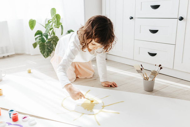 Cute happy little girl, adorable preschooler, painting sun with paints on paper roll sitting on floor in a sunny white living room at home, creative young artist at work - Foto, Bild