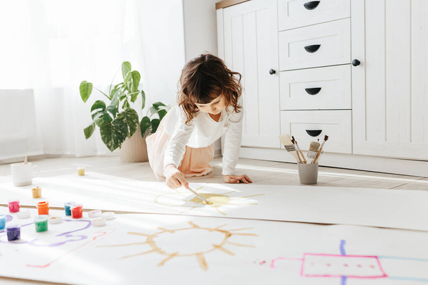 Cute happy little girl, adorable preschooler, painting sun with paints on paper roll sitting on floor in a sunny white living room at home, creative young artist at work - Фото, изображение