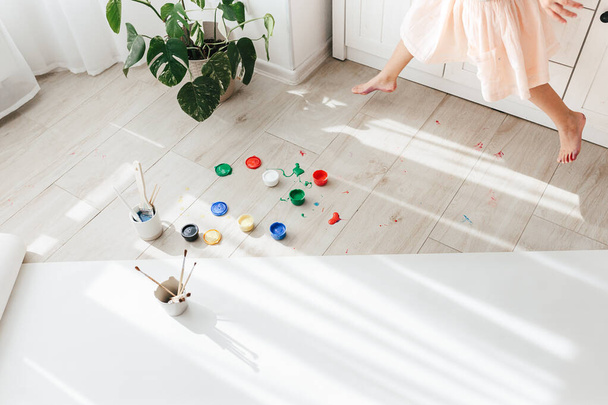 Cute happy little girl, adorable preschooler, painting with paints on paper roll sitting on floor in a sunny white living room at home, creative young artist at work - Фото, изображение