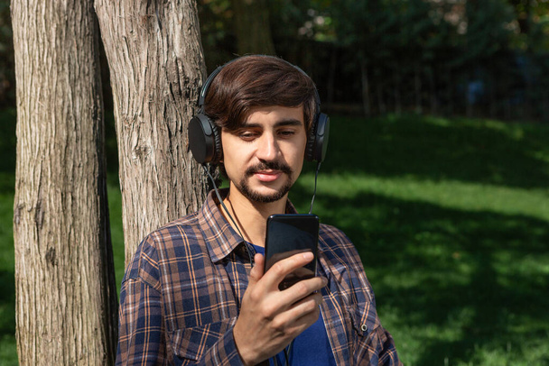 Handsome brunet bearded man in a check shirt listening to music on a smartphone enjoying warm day in a park. - Foto, imagen