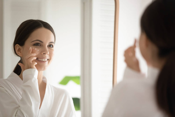 Young woman look in mirror apply facial cream - Foto, Imagen