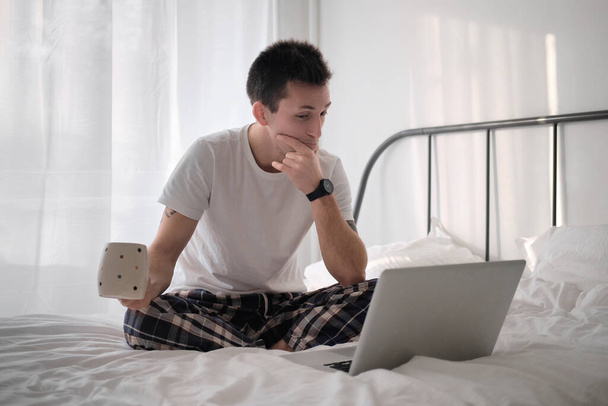Schöner junger Mann im weißen Hemd und Pyjama sitzt in einem Bett mit weißer Bettwäsche und Laptop und trinkt Kaffee und blickt in die Kamera. Arbeiten aus der Ferne während der Isolation. Freiberufler bei der Arbeit. - Foto, Bild