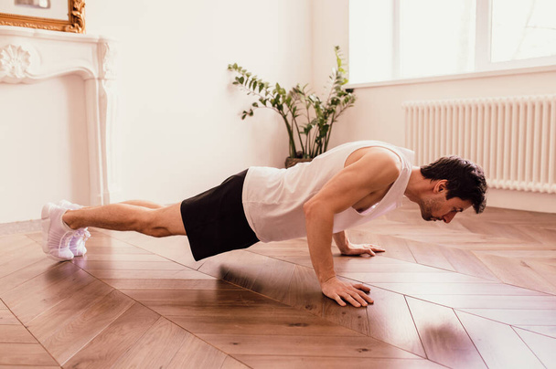  Handsome muscular man in a t-shirt and shorts doing functional exercises on floor at home - Foto, afbeelding