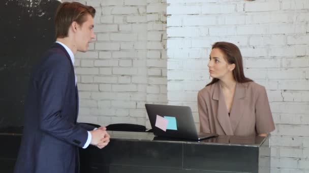 Man in a strict suit approaches a reception in hotel and talks to the girl. Smiling male businessman arrived at hotel having business trip registering on reception - Footage, Video