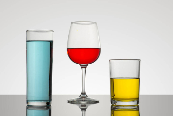 Beker en kristallen glazen met een witte achtergrond en drankjes van verschillende soorten en kleuren, met aantrekkelijke composities - Foto, afbeelding