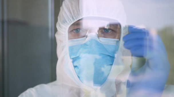 Lääkäri yllään suojapuku, kasvonaamio, lasit ja käsineet COVID-19 eristäminen takana ikkunan sairaalahuoneessa - Materiaali, video