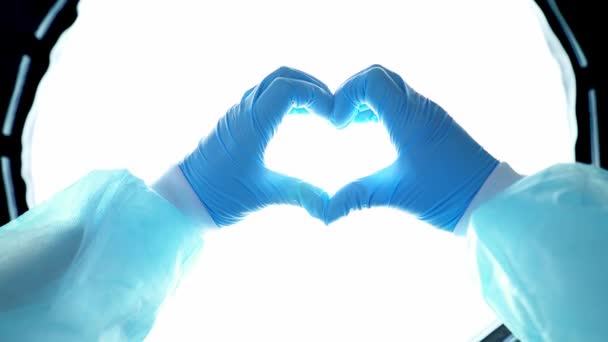 Handen van een arts in blauwe handschoenen maken een hart op de achtergrond van een opererende lamp. Een verpleegster toont een teken van liefde tijdens een pandemie. Dokters redden mensen. COVID-19. Coronavirus - Video