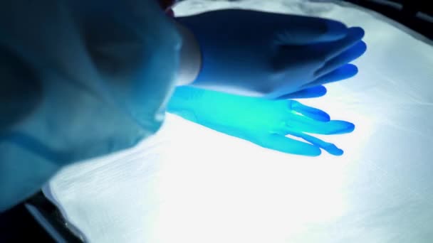 Lekarz zakłada niebieskie rękawice medyczne na sali operacyjnej. Pielęgniarka chroni ręce, by ratować ludzi. Ogromna lampa świeci na dłonie. Chroń ręce przed wirusami. Światowa pandemia, światowy kryzys. - Materiał filmowy, wideo