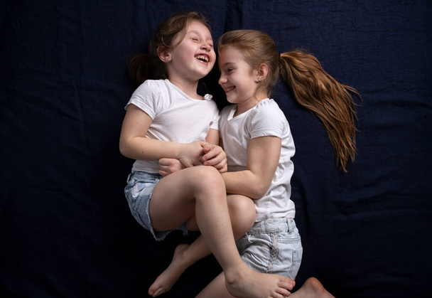 Dziewczyny leżą na niebieskim tle i mają ubaw śmiejąc się łaskocząc się nawzajem. Przyjaźń sióstr. Rozrywka dla dzieci. Zabawa - Zdjęcie, obraz