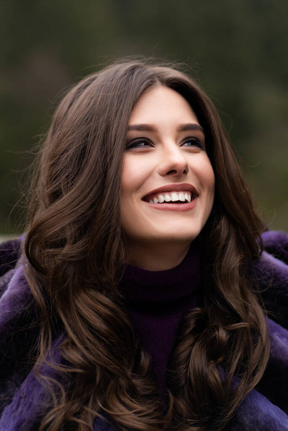 Girl smiling at camera on winter woods background. Glamorous funny young woman with smile wearing stylish sweater, purple fashionable fur coat. Fur and fashion concept. Beautiful people. - Foto, Imagem