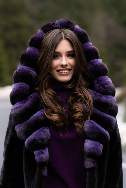 Girl smiling at camera on winter woods background. Glamorous funny young woman with smile wearing stylish sweater, purple fashionable fur coat. Fur and fashion concept. Beautiful people. - Фото, зображення
