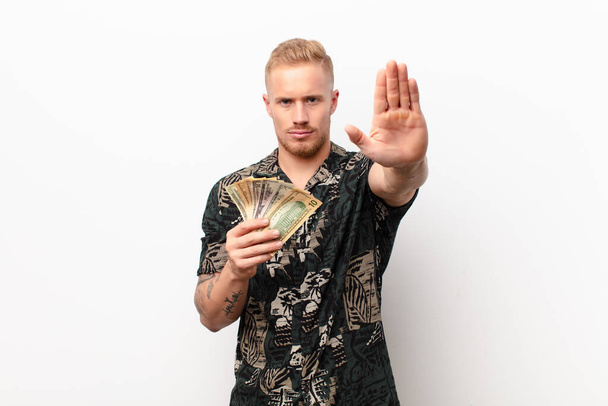 young blonde man looking serious, stern, displeased and angry showing open palm making stop gesture against white wall with banknotes - Fotoğraf, Görsel