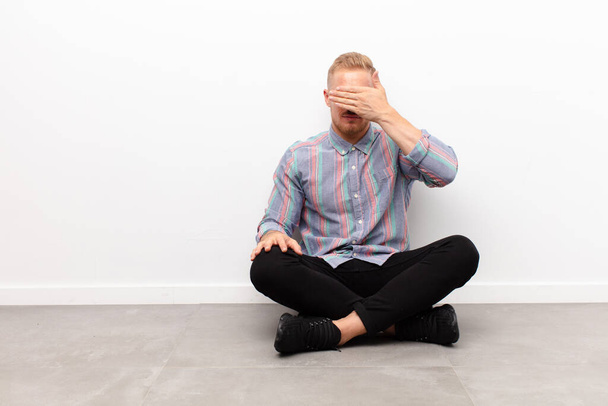 young blonde man covering eyes with one hand feeling scared or anxious, wondering or blindly waiting for a surprise sitting on cement floor - Photo, Image