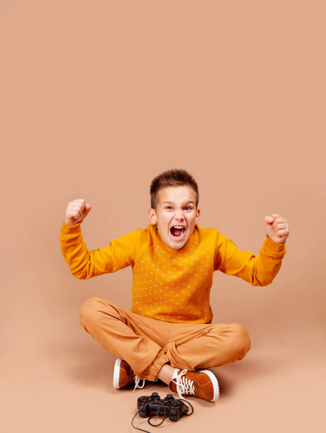 a teenager emotionally plays a computer or console game using the joystick. The boy sits on a beige background - Photo, image