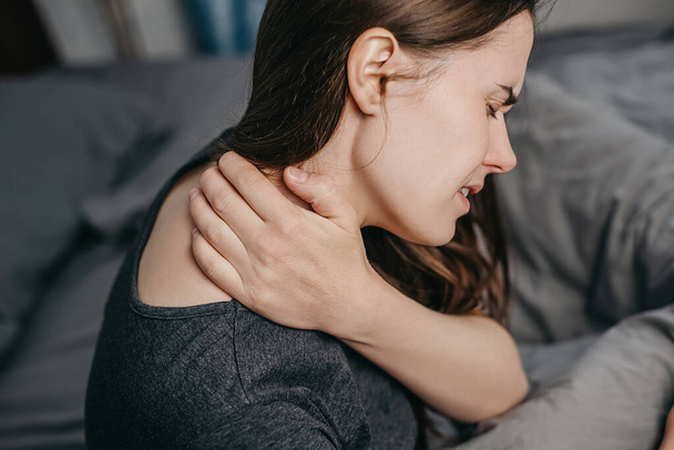 Close up rear view of young woman sitting bed at home scratching her shoulder and neck because of dry skin, dressed in grey t-shirt. Health problem and skin diseases concept - Foto, Bild