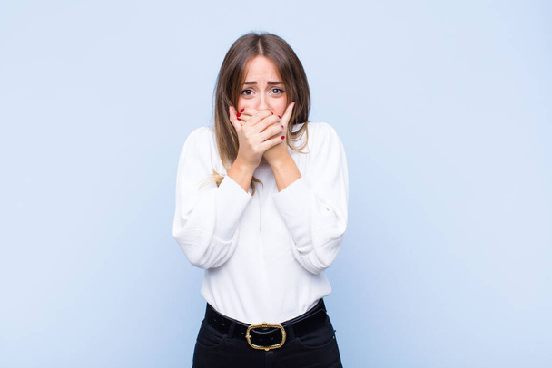 young pretty hispanic woman covering mouth with hands with a shocked, surprised expression, keeping a secret or saying oops against blue wall - Fotoğraf, Görsel