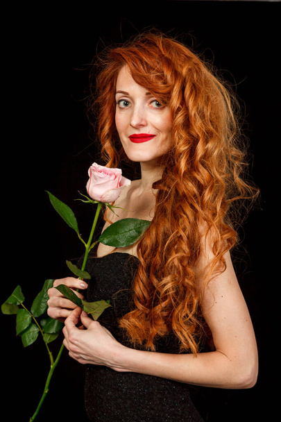 Beautiful girl with red hair in a black dress with a rose on a black background close-up - Fotografie, Obrázek