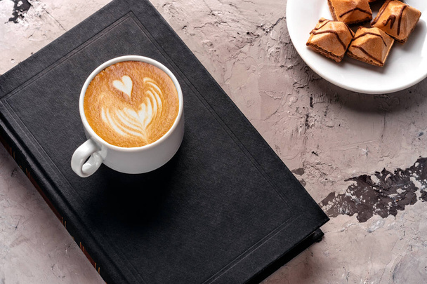 Tasse köstlichen Latte-Kaffee, knusprige Waffeln mit Schokoladenglasur auf einer weißen Untertasse auf grauem Betongrund. Eine Tasse Kaffee steht auf einem schwarzen Buch. Kaffeepause. Beim Kaffee ein Buch lesen - Foto, Bild
