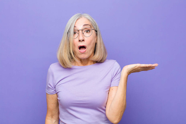 senior or middle age pretty woman looking surprised and shocked, with jaw dropped holding an object with an open hand on the side - Photo, image
