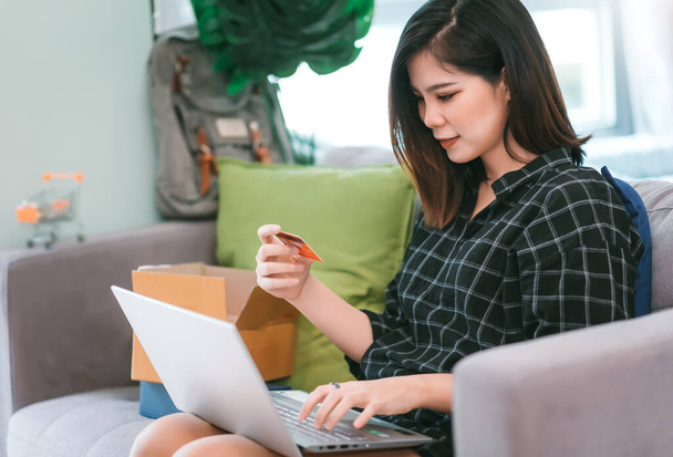 Азиатская молодая женщина держит кредитную карту и с помощью ноутбука для покупок в Интернете на дому гостиной - Работа из дома и остаться дома концепции
 - Фото, изображение
