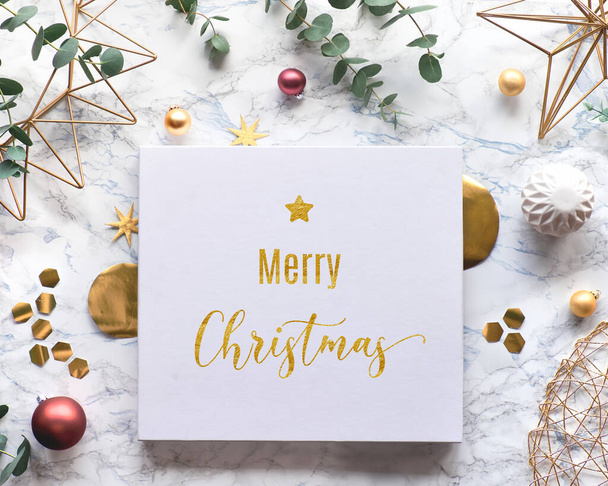 Gilded text "Merry Christmas". Christmas frame with fresh eucalyptus twigs and golden geometric decorations - hexagons and metal wire shapes. Trendy flat lay, top view on white marble background. - Photo, image