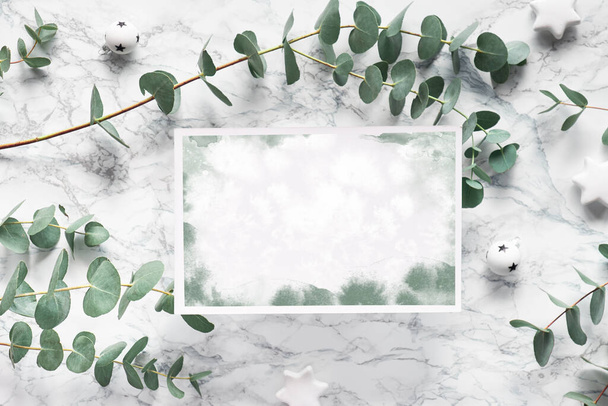 Christmas frame with fresh eucalyptus twigs and white trinkets with black stars. Flat lay marble background. Paper page with watercolor pattern, copy-space, text place. - Fotografie, Obrázek