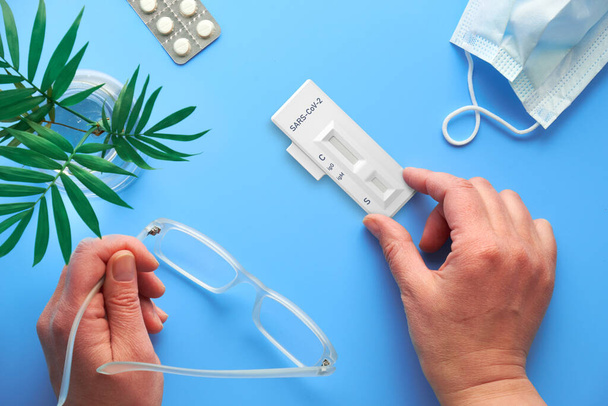 Rapid COVID-19 test in hand. Serological detection of antibodies IgM and IgG to novel coronavirus SARS-CoV-2 causing Covid-19. Blue background with palm plant, reading glasses, pills and face mask. - Valokuva, kuva