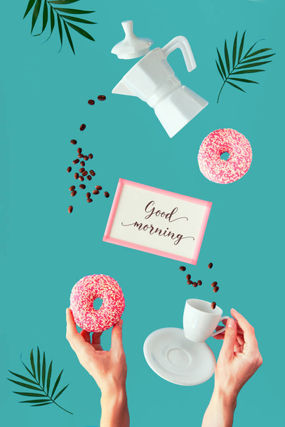 Levitation coffee and doughnut in hands. Flying line of coffee beans. ceramic coffee maker and espresso cup held by hand. Palm leaves, frame with text "Good morning". Vibrant green mint background. - Photo, image