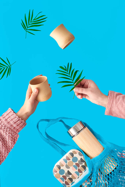 Surreal levitation on vibrant blue mint background. Two hands with bamboo cup and palm leaf. Zero waste tea in in eco friendly insulated bamboo flask. Net.bag with lunch box and tea flask. - Foto, afbeelding
