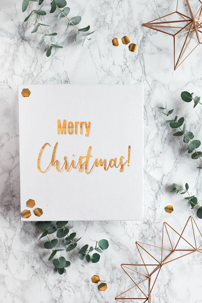 Gilded text "Merry Christmas". Christmas frame with fresh eucalyptus twigs and golden geometric decorations - hexagons and metal wire shapes. Flat lay, top view on white marble background. - Foto, Bild