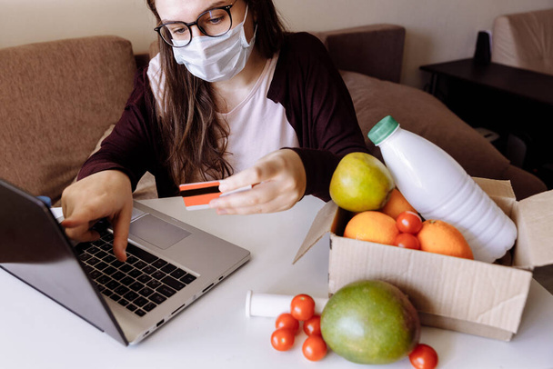 A girl in a gauze mask and glasses makes an order in an online store. On white table is a laptop and box of vegetables.  - Fotó, kép