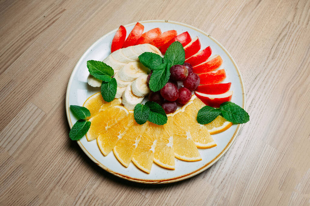 φρούτα φέτες μπανάνα μήλο πορτοκάλι και το σταφύλι με φύλλα δυόσμου σε ένα λευκό παγωμένο πιάτο σε ξύλινο τραπέζι φόντο - Φωτογραφία, εικόνα