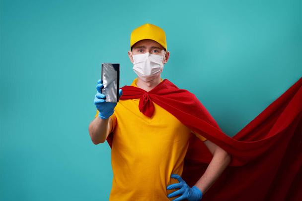 Mensajero vestido con capa de superhéroe y máscara médica sostiene el teléfono inteligente en su mano
. - Foto, Imagen